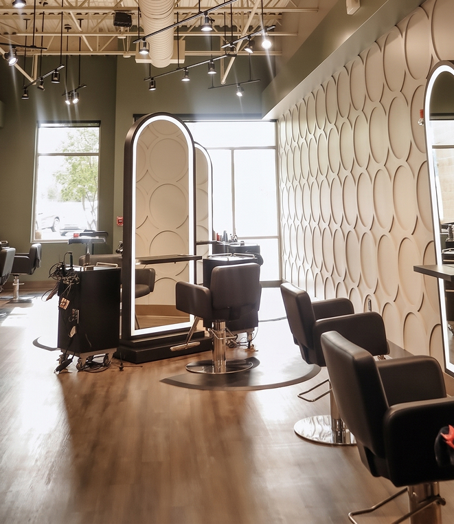 Hair salon interior with several black styling chairs facing oval LED-lit mirrors against a textured wall. - Salon Aria + Spa in Davenport, IA